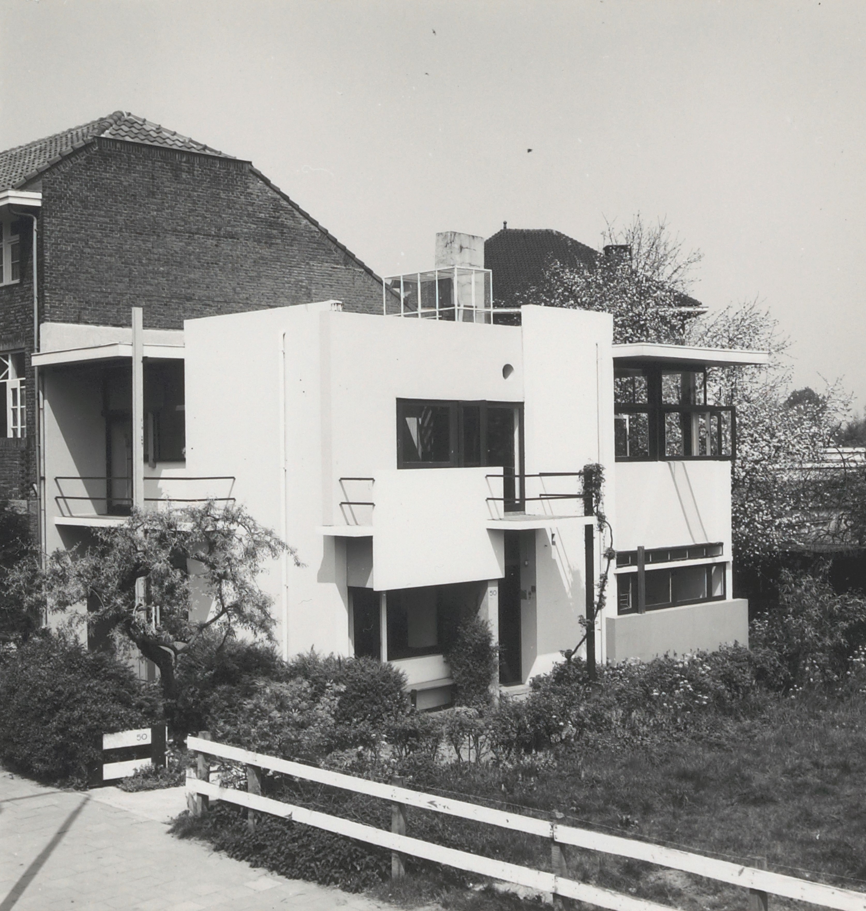 De foto toont voornamelijk de voorkant, en een deel van de zijkant van het huis. De tuin is begroeit met struiken en hoog gras. 